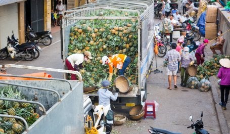 Three people unload pineapple from a truck on a busy street in Vietnam.